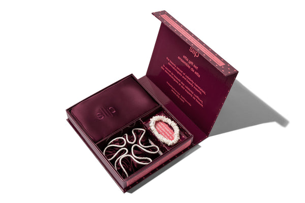 An open gift set box containing a silk crimson pillowcase, a crimson large scrunchie and a white skinny silk scrunchie, photographed on a clean white background.