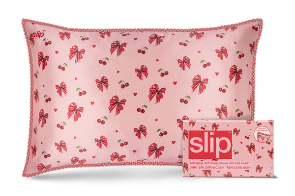 A pink Slip brand silk pillow with a red bow and cherry pattern and lace trim, shown next to its matching pink packaging box on a white background.