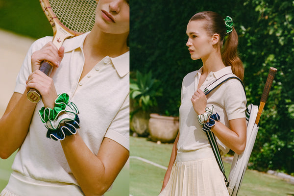 Woman holding a vintage tennis racket on a tennis court with large silk scrunchies on her wrist and greenery in the background